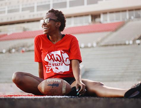 A female 欧洲杯买球app student wearing a Jax State tee sits on the field at 欧洲杯买球app stadium with a football in front of her